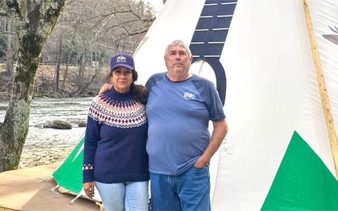 SMT photo/Sara Eisinger.  Grumpy Bear Campground owners Jerry and Karen Ciullo stand outside six brand new 18-foot-tall teepees along Tuckasegee River Monday, March 24. Hurricane Helene flooded the campground last September, taking its teepees down river. 